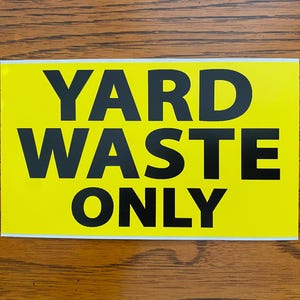 May include: A rectangular sign with a bright yellow background and bold black text that reads "YARD WASTE ONLY". The sign has a white border and is displayed on a wooden surface.