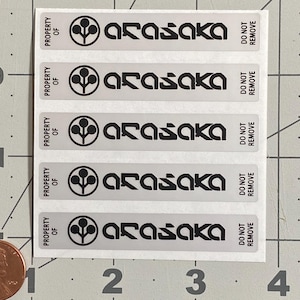 May include: A sheet of rectangular stickers with the text "PROPERTY OF ARASAKA DO NOT REMOVE" in black on a silver background. Each sticker features a stylized logo. A penny is in the lower left corner for scale.