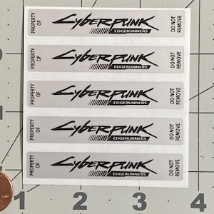 Puede incluir: Una hoja de pegatinas rectangulares plateadas con el texto "CYBERPUNK EDGERUNNERS" en negro. Las pegatinas tambi&eacute;n dicen "PROPERTY OF" y "DO NOT REMOVE". Una moneda est&aacute; en la esquina inferior izquierda para la escala.