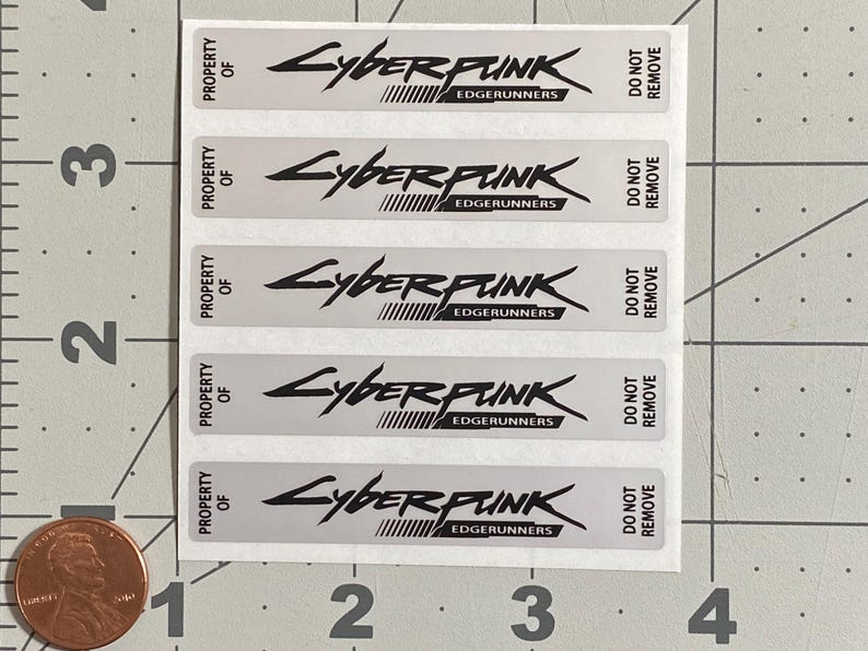 Puede incluir: Una hoja de pegatinas rectangulares plateadas con el texto "CYBERPUNK EDGERUNNERS" en negro. Las pegatinas tambi&eacute;n dicen "PROPERTY OF" y "DO NOT REMOVE". Una moneda est&aacute; en la esquina inferior izquierda para la escala.