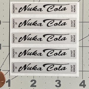 May include: A sheet of five rectangular stickers with the text "Nuka Cola" in a stylized black font. Each sticker has the words "PROPERTY OF" and "DO NOT REMOVE" printed on it. The stickers are on a white background.