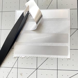 May include: A sheet of white rectangular labels with a silver, peeling top corner. Black tweezers are lifting the corner of the label, revealing the adhesive. The labels are on a gray cutting mat with grid lines and numbers.