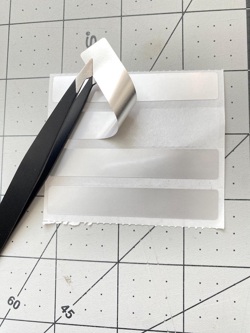 May include: A sheet of white rectangular labels with a silver, peeling top corner. Black tweezers are lifting the corner of the label, revealing the adhesive. The labels are on a gray cutting mat with grid lines and numbers.