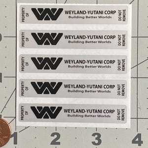 May include: A sheet of five rectangular stickers. Each sticker has the text "PROPERTY OF" above a black "W" logo, followed by "WEYLAND-YUTANI CORP Building Better Worlds" and "DO NOT REMOVE". The stickers are on a grid background.