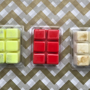May include: Three wax melts in clear plastic packaging. The wax melts are in the shape of a rectangle and are divided into six squares. The wax melts are yellow, red, and white.