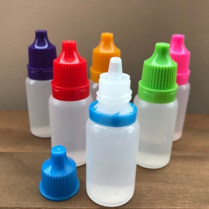 May include: Six clear plastic bottles with colourful dropper caps. The bottles are arranged in a semi-circle on a wooden surface. The caps are purple, red, orange, green, pink, and blue.