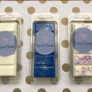 May include: Three wax melts in clear plastic packaging. The wax melts are in the scents: white, blue with glitter, and white with purple glitter. Each wax melt has a round sticker with a pink heart and the text "Cosy up Candles".