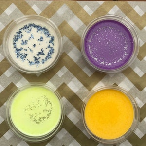 May include: Four small glass containers filled with different coloured wax melts. The wax melts are white with blue glitter, purple with silver glitter, green with gold glitter, and yellow with gold glitter.