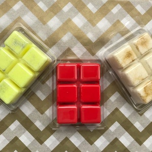 May include: Three packs of scented wax melts in clear plastic packaging. The melts are yellow, red, and off-white. The yellow melts have silver glitter, the red melts are solid, and the off-white melts have gold glitter. The background is a gold and white chevron pattern.