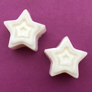 May include: Two white star-shaped wax melts with a layered design, each with a smaller star shape inside. The melts are on a deep purple background, creating a contrast that highlights the star shapes. These are likely for use in a wax warmer.