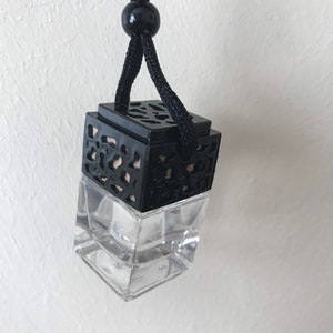May include: A car air freshener with a clear glass bottle and a black, decorative top. The top features an openwork geometric design. A black cord and bead are attached for hanging. The freshener hangs from a black cord.