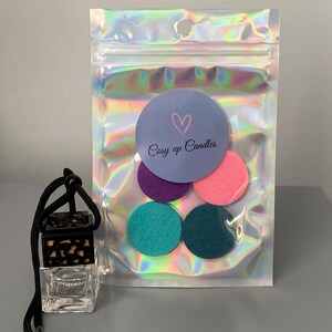 May include: A clear glass car air freshener with a black and brown patterned lid, hanging from a black string. The air freshener is packaged with a small bag of five felt scent discs in purple, pink, teal, and dark blue.