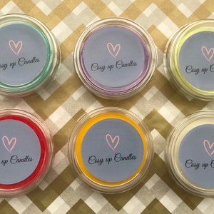 May include: Six small glass jars with lids, each containing a wax melt. The wax melts are in various colours, including green, purple, yellow, red, orange, and white. Each jar has a label with the text "Cosy up Candles" and a pink heart.