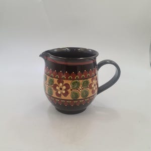 May include: A dark brown ceramic pitcher with a handle and spout. The pitcher is decorated with a floral pattern in shades of red, yellow, and green. The rim and handle are black.