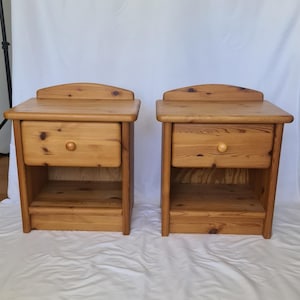 Op de afbeelding: Een paar bijpassende nachtkastjes van lichtbruin hout, elk voorzien van een enkele lade met een ronde houten knop, een plat bovenblad met een gebogen achterwand en een open opbergplank direct onder de lade.
