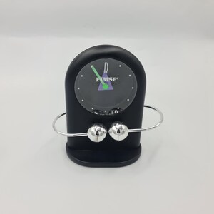 May include: A black desk clock with a silver pendulum and two silver spheres. The clock face is dark gray with a green and purple triangle and the word "FIMSE". The clock has a curved silver bar that holds the spheres.