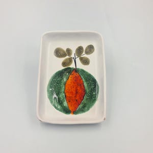 May include: Rectangular ceramic dish with a hand-painted design. The design features a green and orange fruit with a small branch and leaves. The dish is white with a slightly raised edge.