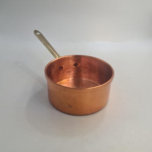 Puede incluir: Una pequeña cacerola de cobre con un mango de latón. La cacerola tiene un fondo redondeado y un borde ligeramente acampanado.