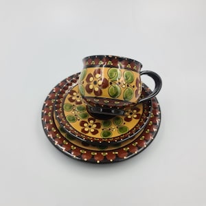 May include: A ceramic tea set with a floral design. The set includes a mug and two plates. The mug and plates have a yellow background with brown and green floral patterns. The rims are brown with white dots.