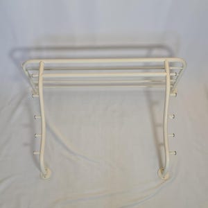 May include: A white metal wall-mounted rack featuring a top shelf with three horizontal bars and two curved side supports. Each side support includes three small hooks extending outward, providing a total of six hooks for hanging items.