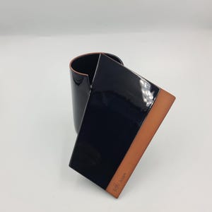 May include: A modern, abstract ceramic vase with a glossy black exterior and a contrasting tan-colored edge. The vase has a unique, curved design with a rectangular panel. The artist's signature is visible on the tan edge.