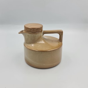 May include: A cylindrical ceramic pitcher with a light tan, speckled glaze and a smooth, darker brown lid. The vessel features a short, integrated spout and a flat, angular handle extending from the side, showcasing a uniform, glossy finish.