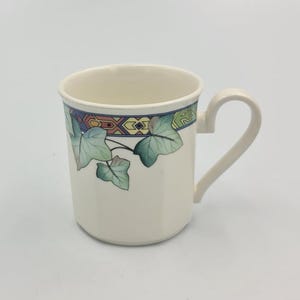 Könnte beinhalten: Cremefarbene Keramiktasse mit gebogenem Henkel. Die Tasse hat ein dekoratives Band aus geometrischen Formen in Blau, Orange und Grün am Rand, mit grünen Efeublättern an der Seite. Klassisches Design.