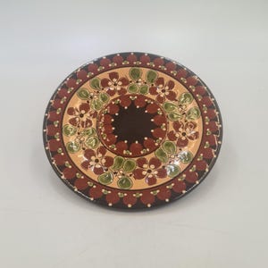 May include: A round, decorative ceramic plate with a detailed floral design. The plate features a dark brown center, surrounded by a tan band with green and white floral patterns. The outer rim is a dark brown with red and white accents.