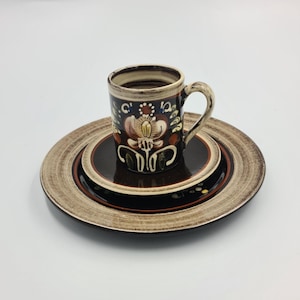 Op de afbeelding: Een keramische koffiekop met een bloemmotief in bruin, beige en rood staat op een bijpassende set borden. De borden hebben een bruin en beige gestreept patroon. De kop en borden vormen een set.