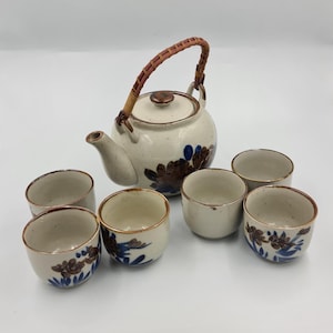May include: A ceramic tea set comprising a teapot with a woven handle and lid, and six matching teacups. The set is off-white with brown and blue floral designs. The teapot has a spout and a rounded body.