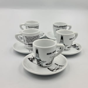 May include: Six white espresso cups and saucers. Each cup features a cartoon character and the words "MOON EXPRESSO" in black. The saucers also have the same cartoon character and text. The cups and saucers are arranged on a white surface.