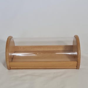 Könnte beinhalten: Ein Brotkasten aus Holz mit einem durchsichtigen, gebogenen Kunststoffdeckel. Der Kasten besteht aus hellfarbenem Holz und hat eine rechteckige Basis. Der transparente Deckel ermöglicht die Sichtbarkeit des Inhalts. Das Design ist einfach und funktional.