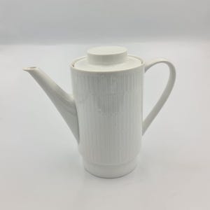 Könnte beinhalten: Eine weiße Kaffeekanne aus Keramik mit Deckel und Griff. Die Kanne hat einen zylindrischen Körper mit einem strukturierten, vertikalen Streifenmuster. Der Ausguss ist lang und abgewinkelt. Der Griff ist gebogen. Die Kanne steht auf weißem Hintergrund.
