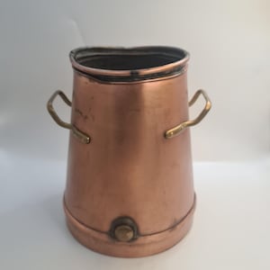 May include: A copper-coloured metal container with a flared shape, featuring two brass handles and a brass knob near the base. The container has a dark rim, likely vintage or antique.