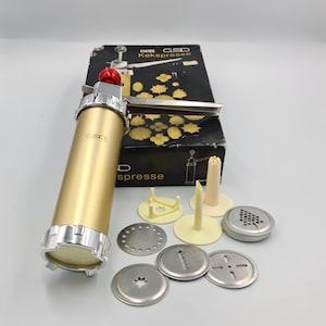 May include: A gold-colored cookie press with a red top and multiple stainless steel cookie shape discs. The box reads "EMIDE GSD Kekspresse". Includes various nozzles and a box with cookie shape illustrations.