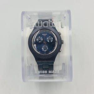 May include: A dark blue Swatch watch in a clear plastic case. The watch has a dark blue face with white numerals and hands. The watch band and case are also dark blue. The text "SWISS MADE" is visible.