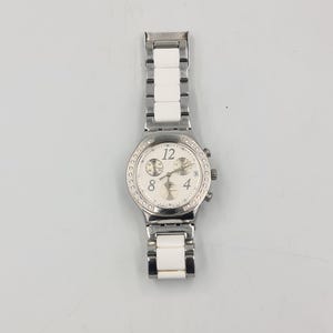 May include: A silver-toned wristwatch with a white face and a bezel encrusted with small, clear stones. The watch features a white and silver band with rectangular links. The watch face displays the numbers 12, 4, and 8, along with smaller dials and hands.