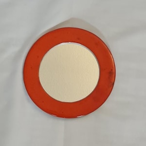May include: A round, orange compact mirror with a silver-colored reflective surface. The mirror is surrounded by a bright orange frame. The compact is a simple, yet stylish accessory.