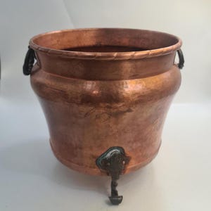 May include: A copper-coloured planter with a wide, rounded body and a textured rim. It has two black handles and a decorative foot. The planter is likely made of copper and has a vintage or antique appearance.