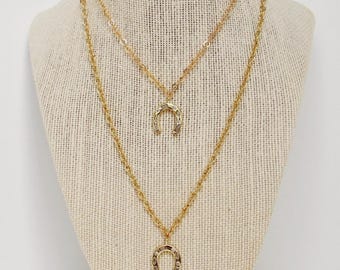 May include: Two gold-colored necklaces with horseshoe pendants displayed on a mannequin. The necklaces are layered, with one pendant slightly smaller than the other. The chains are delicate, and the pendants are detailed.