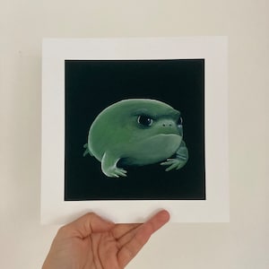 May include: A green frog painting on a black background, presented on white matted paper. The frog has a grumpy expression and is the focal point of the artwork. The artwork is held by a hand.