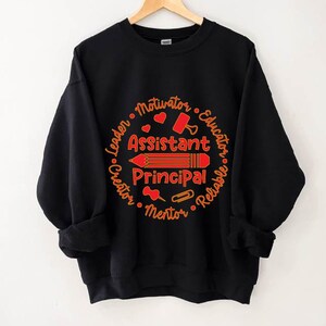 May include: Black sweatshirt with a red and orange design. The design features the words "Assistant Principal" with a pencil illustration, surrounded by words like "Leader" and "Educator". A perfect gift for school staff.