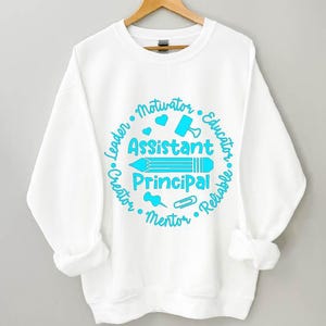 May include: White sweatshirt with a crew neck and long sleeves. The front features a teal graphic with the words "Assistant Principal" and surrounding words like "Leader", "Educator", and "Mentor". A pencil, heart, and other school-related icons are included.