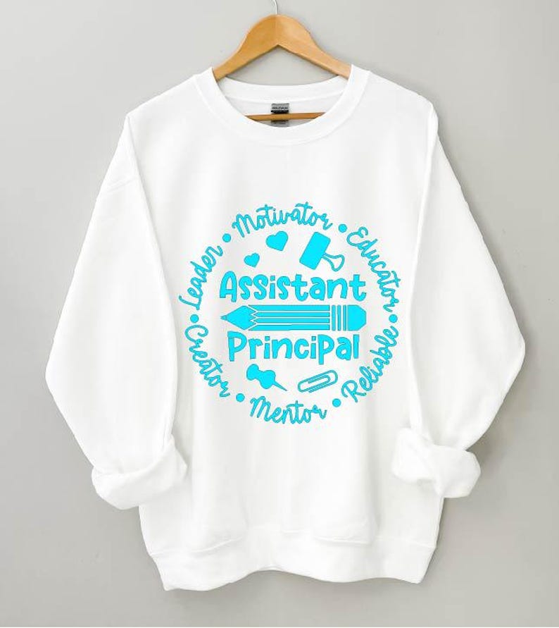 May include: White sweatshirt with a crew neck and long sleeves. The front features a teal graphic with the words "Assistant Principal" and surrounding words like "Leader", "Educator", and "Mentor". A pencil, heart, and other school-related icons are included.