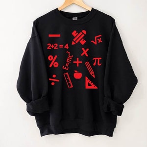 Puede incluir: Sudadera negra con s&iacute;mbolos matem&aacute;ticos rojos y &uacute;tiles escolares impresos en ella. Los s&iacute;mbolos incluyen un signo m&aacute;s, un signo menos, un signo de multiplicaci&oacute;n, un signo de divisi&oacute;n, un signo de porcentaje, pi, una ra&iacute;z cuadrada y la ecuaci&oacute;n E=mc2. Los &uacute;tiles escolares incluyen un l&aacute;piz, una regla, un transportador y una manzana.
