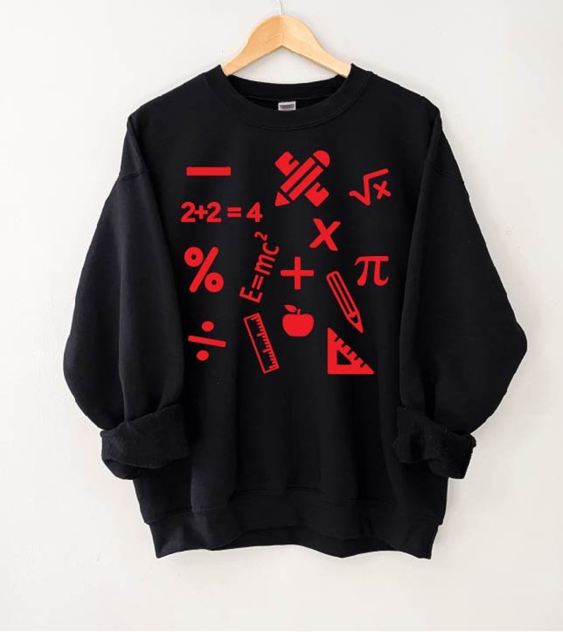 Puede incluir: Sudadera negra con s&iacute;mbolos matem&aacute;ticos rojos y &uacute;tiles escolares impresos en ella. Los s&iacute;mbolos incluyen un signo m&aacute;s, un signo menos, un signo de multiplicaci&oacute;n, un signo de divisi&oacute;n, un signo de porcentaje, pi, una ra&iacute;z cuadrada y la ecuaci&oacute;n E=mc2. Los &uacute;tiles escolares incluyen un l&aacute;piz, una regla, un transportador y una manzana.