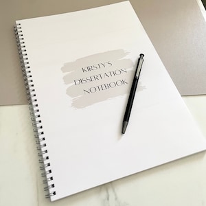 May include: A white spiral bound notebook with the text "Kirsty's Dissertation Notebook" on the front cover. A black pen is lying on the notebook.