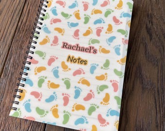 May include: Spiral-bound notebook with a white cover patterned with colorful baby footprints. The notebook has the text "Rachael's Notes" in pink and yellow. The pages are lined.