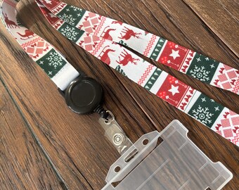 May include: A Christmas-themed lanyard with a festive pattern of reindeer, snowflakes, and stars in red and green on a white background. The lanyard includes a black retractable reel and a clear plastic ID holder.