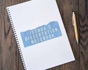 May include: A white spiral bound notebook with a blue and white torn paper design. The text "Joshua's Dissertation Notebook" is printed in white on the blue paper.